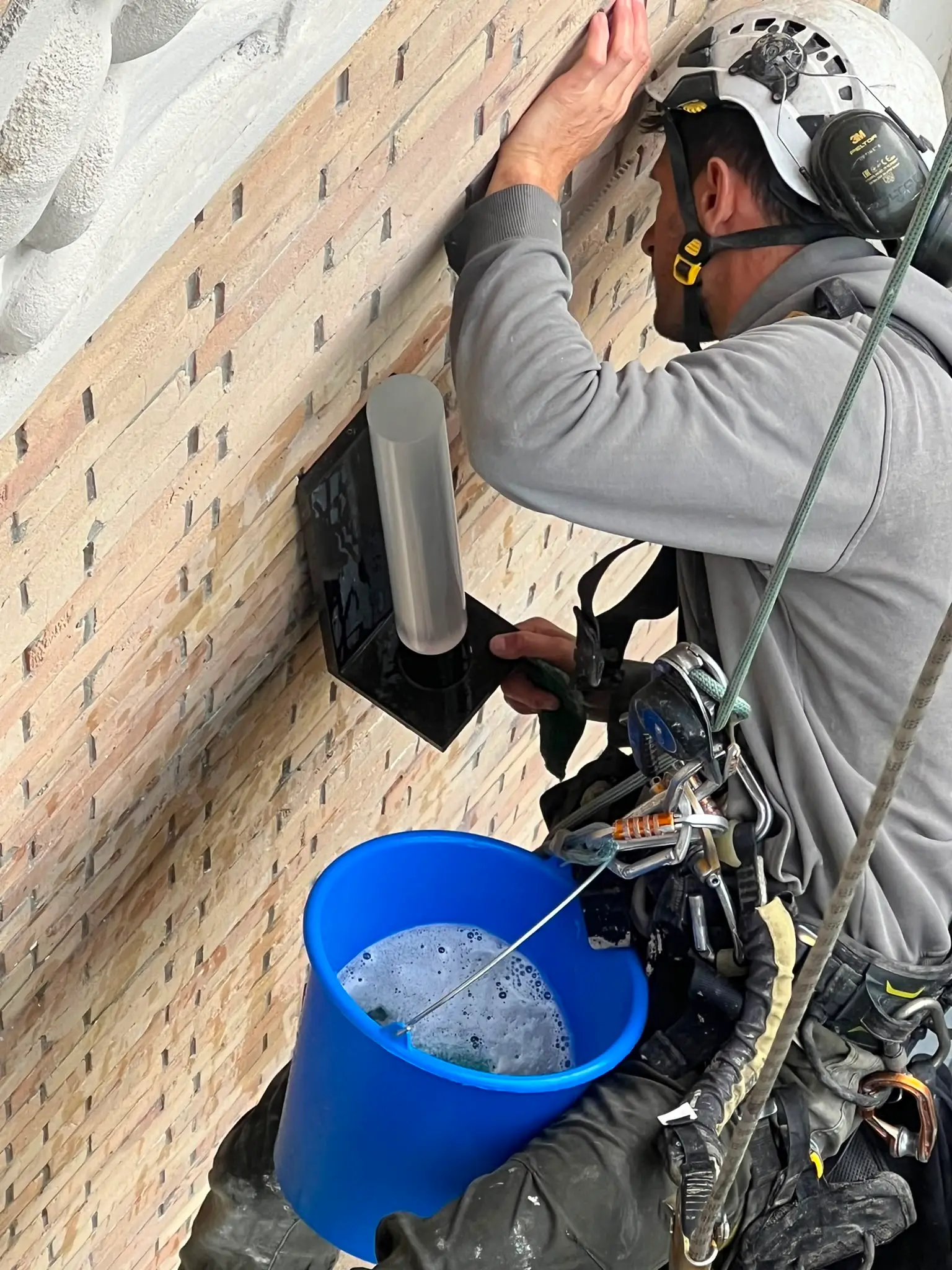 Cordiste casqué, en suspension sur ses cordes, nettoyant à l'éponge un luminaire en inox fixé sur une façade en brique