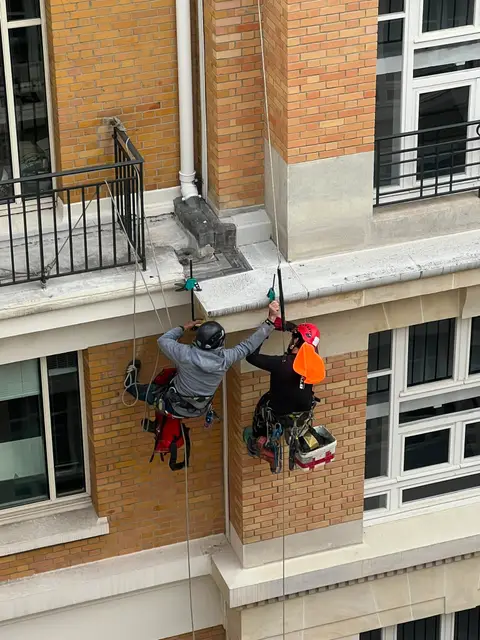 Deux cordistes suspendus côte à côte, grattant le dessous d'un bandeau en pierre sur une façade parisienne en brique