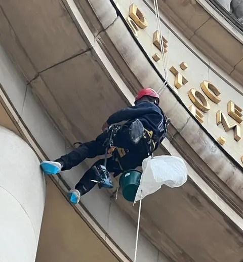 Cordiste au casque rouge suspendu à ses cordes devant la façade en pierre d'un édifice monumental, travaillant sous une frise de lettres dorées