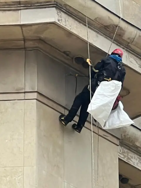 Cordiste suspendu à l'angle d'un immeuble en pierre de taille, nettoyant une zone encrassée sous la corniche, sac de chantier accroché au harnais