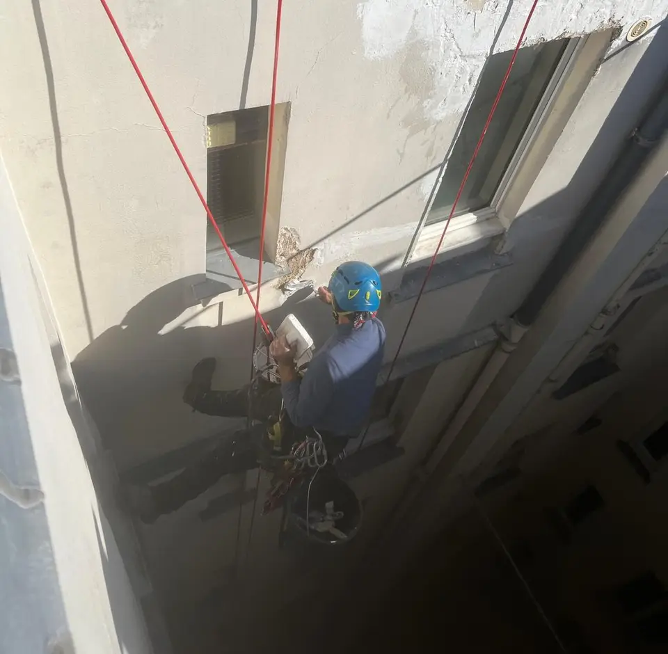 Cordiste au casque bleu, suspendu à ses cordes rouges, reprenant l'enduit d'une façade claire, vu en plongée