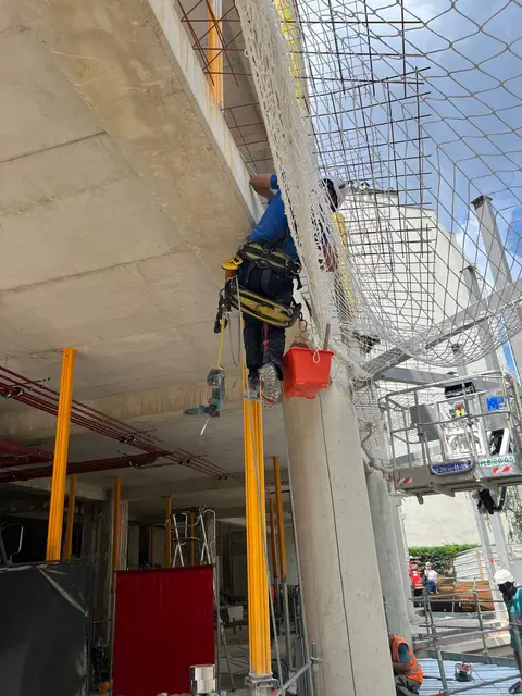 Cordiste suspendu sous une dalle béton, fixant un filet de protection le long du nez de dalle, seau de matériel accroché au harnais