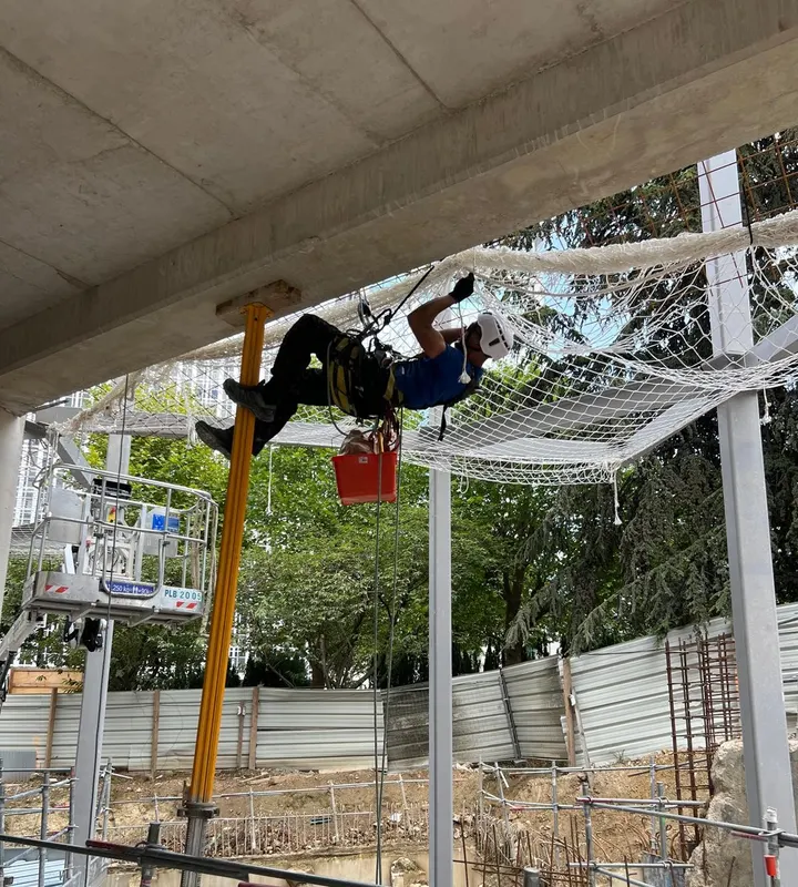 Filet de sécurité horizontal tendu sous une charpente en bois, vu en contre-plongée sur ciel bleu