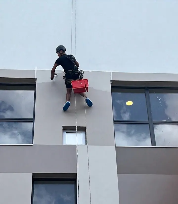 Cordiste en rappel appliquant de la peinture sur la façade supérieure d'un bâtiment moderne, bac de peinture rouge suspendu au harnais