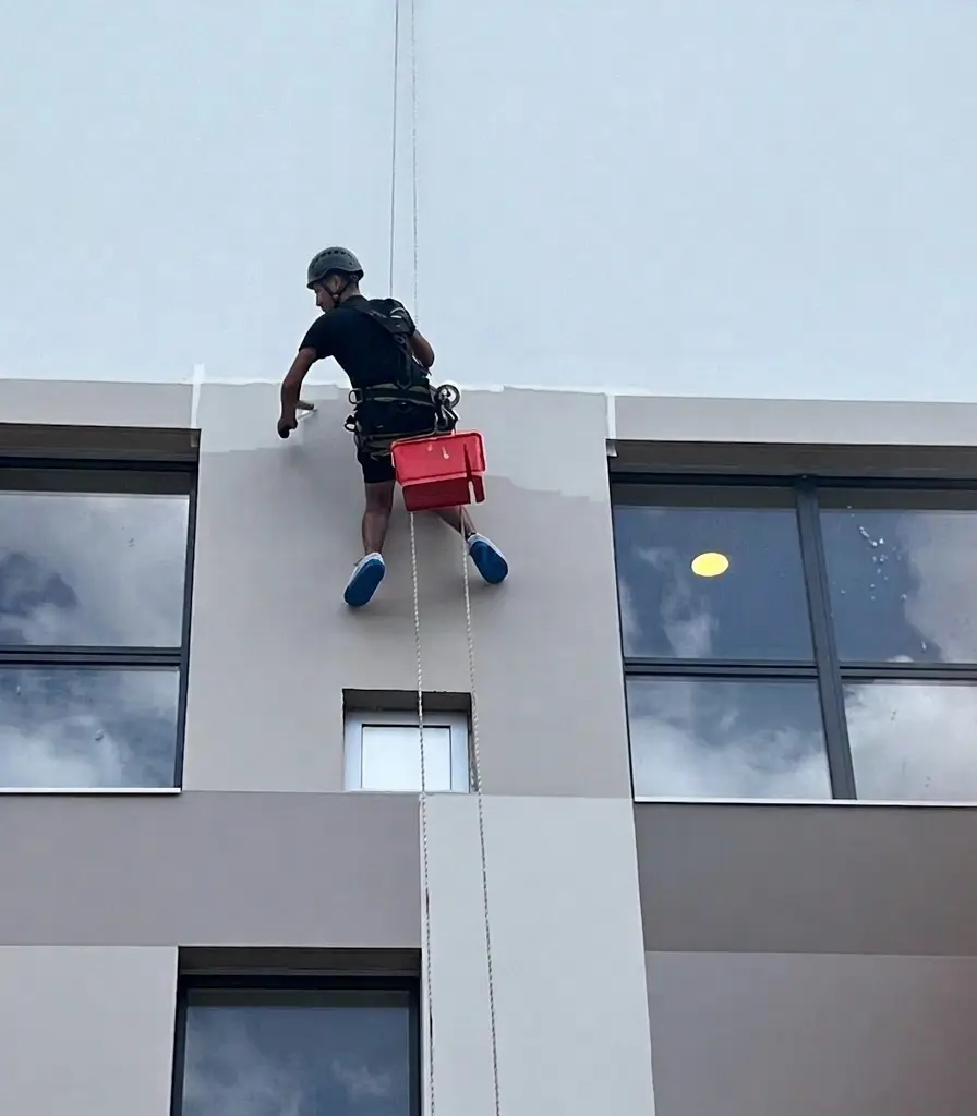 Cordiste en rappel appliquant de la peinture sur la façade supérieure d'un bâtiment moderne, bac de peinture rouge suspendu au harnais